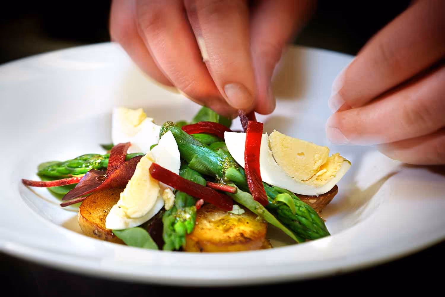 A close up photo of a chef adding the final touches to a scallop, egg, and asparagus dish