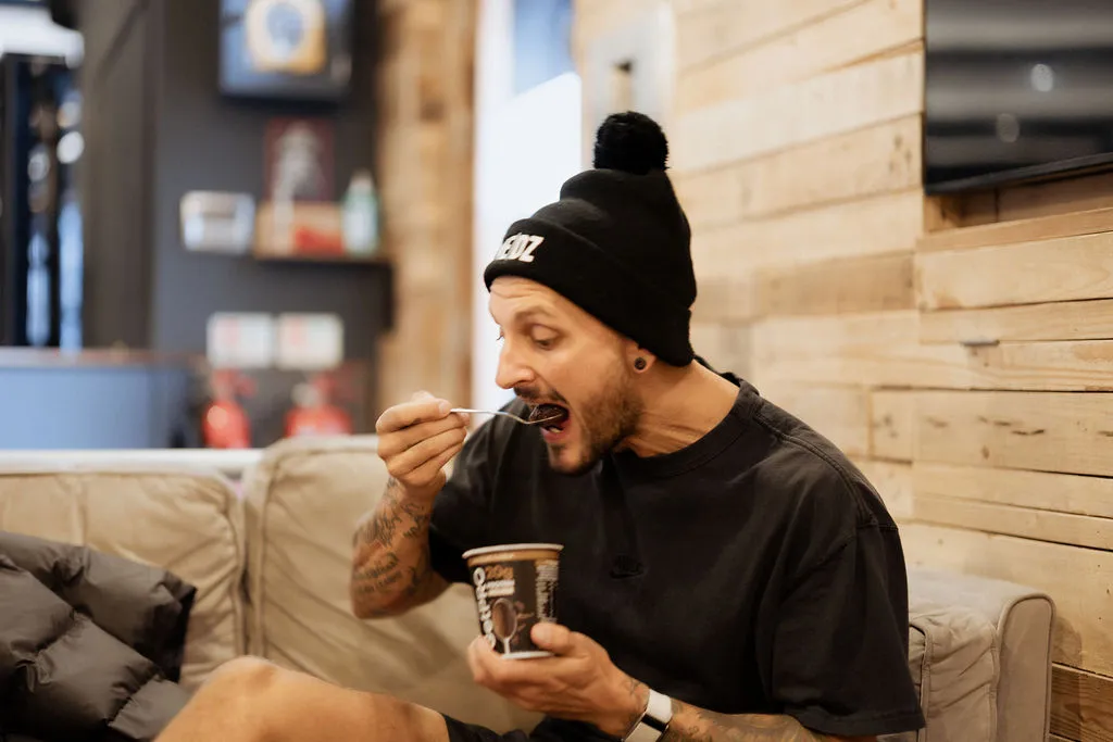 Man wearing a black beanie and T-shirt sitting on a couch and eating from a cup with a spoon in a wooden-paneled room.