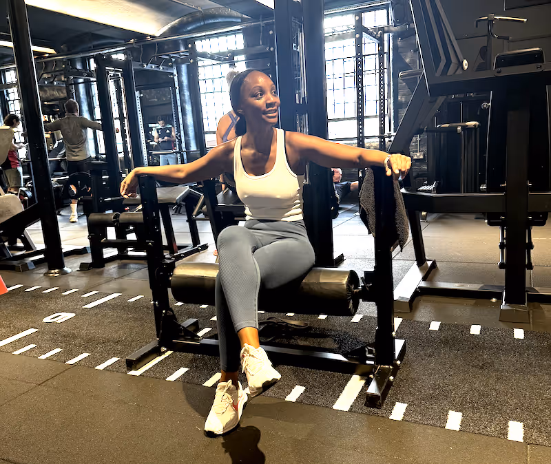 Member sitting on the leg curl machine inside ONE|DZ Gym in Derby, smiling post-workout in a light-filled strength area