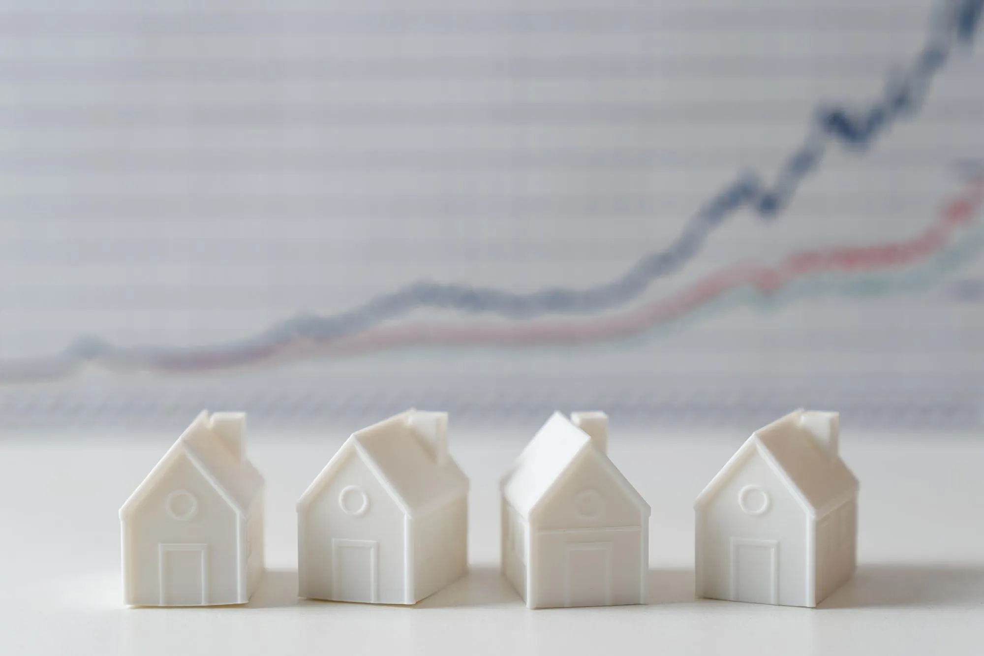Vier weiße Miniaturhäuser vor einem unscharfen Preischart symbolisieren die Entwicklung und Bewertung von Immobilienwerten.