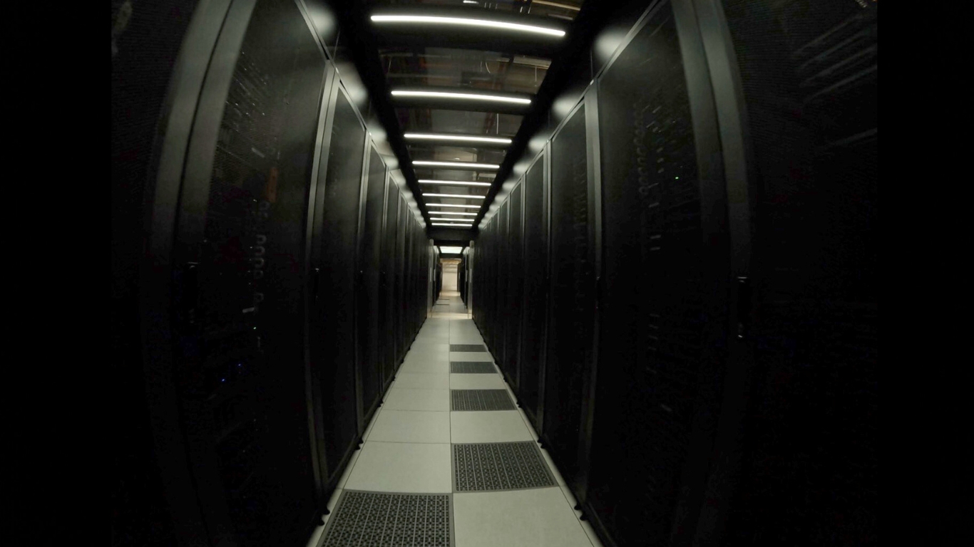 Long corridor between rows of black server racks in a data center with overhead lighting.