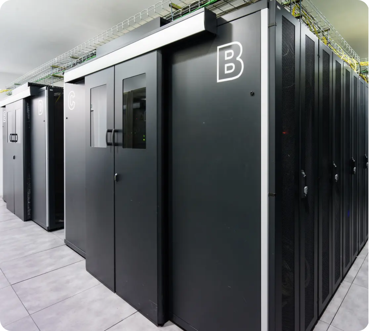 Rows of black server racks labeled B, C, and D in a data center with tiled floor and cable trays overhead.