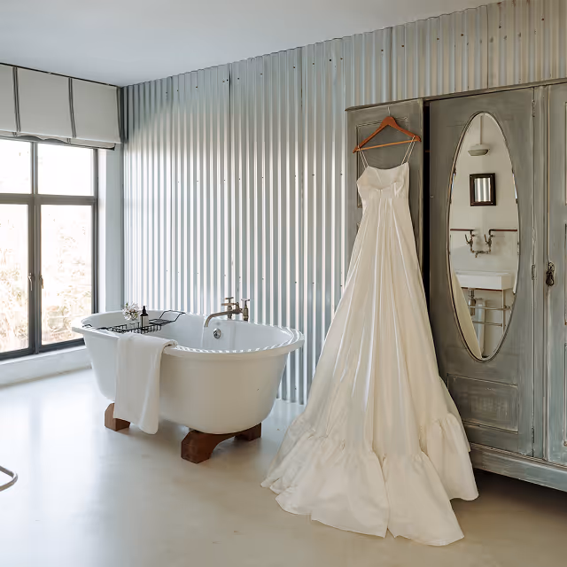 White wedding dress hanging on a rustic gray wardrobe with an oval mirror, next to a white freestanding bathtub in a bright bathroom with corrugated metal walls.