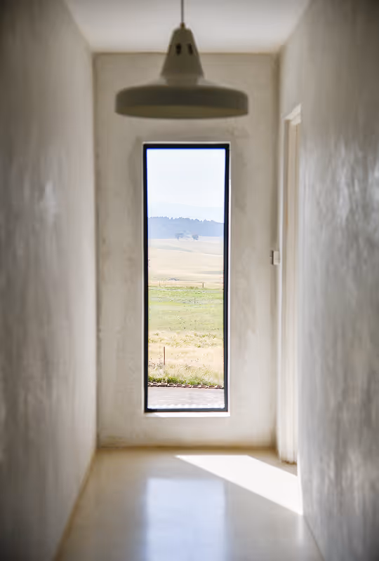 Narrow hallway with white walls and floor lighted by sunlight through a tall, slim window showing a grassy field and distant hills.