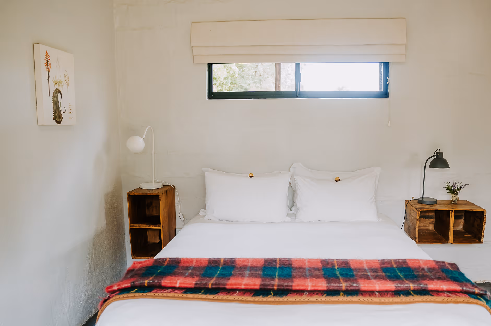 Minimalist bedroom corner with white bed, two pillows, red and green plaid blanket, wooden nightstand, white lamp, small window, and botanical wall art.