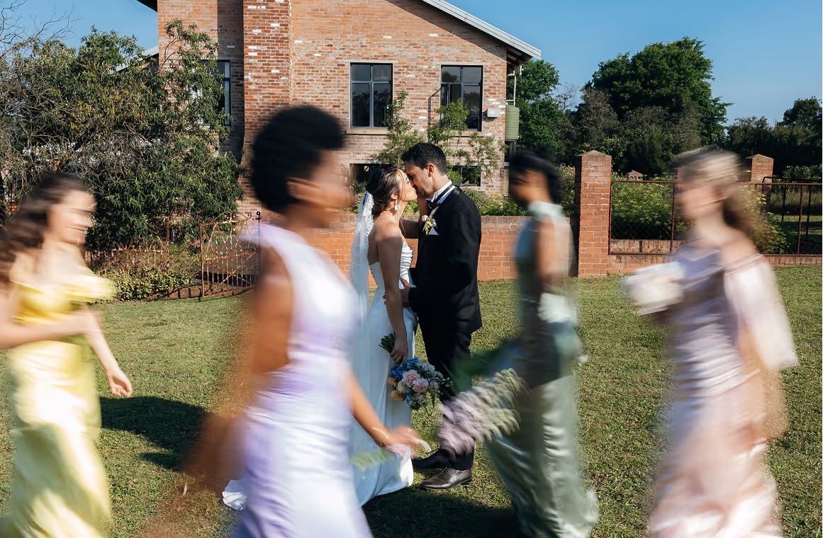 Bride and groom kissing outdoors with bridesmaids in colorful dresses walking blurred past them on a sunny day.