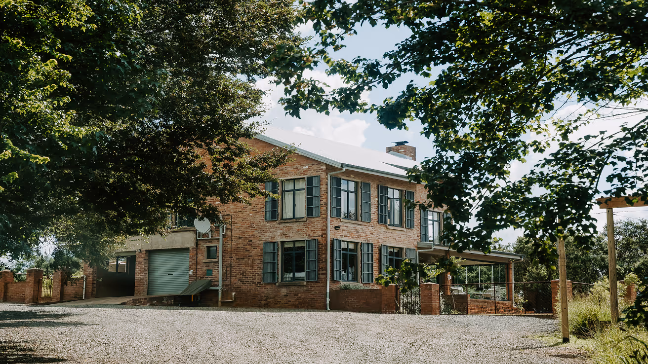 Two-story brick house with multiple windows and a garage, partially framed by leafy tree branches on a sunny day.