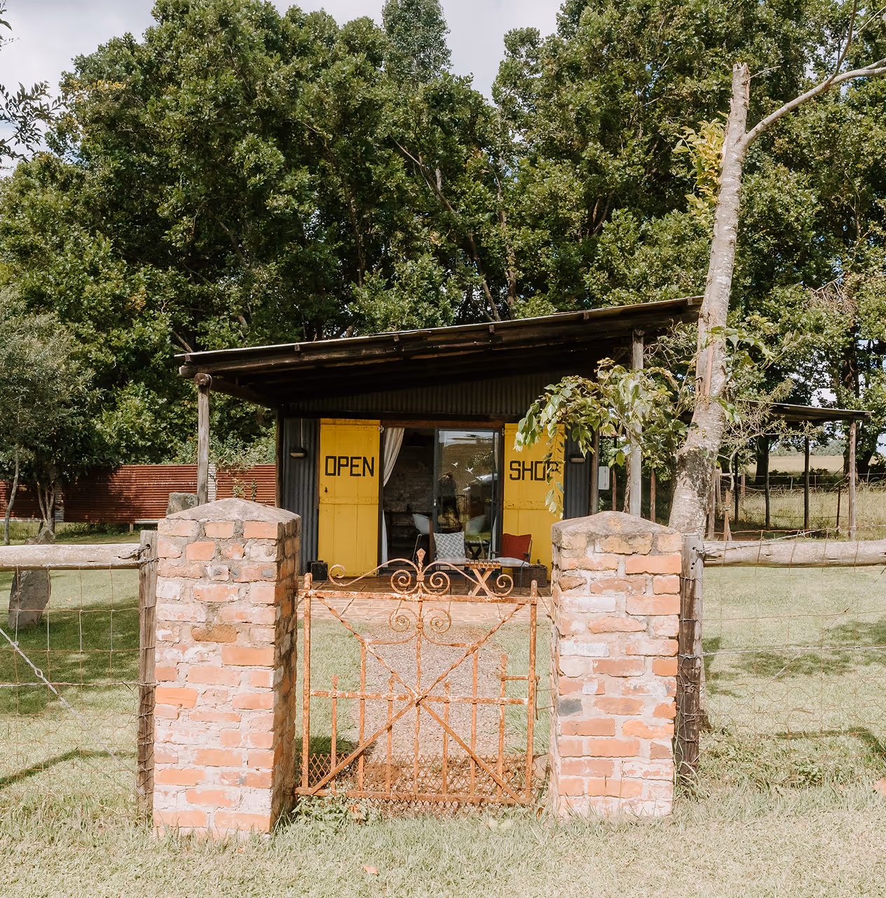 Small rustic shop with yellow doors labeled 'OPEN SHOP' behind a rusty metal gate and brick pillars, surrounded by green grass and trees.