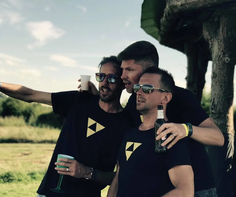 Three men wearing matching Beat Bunker shirts posing outdoors with drinks, smiling and pointing in the distance.
