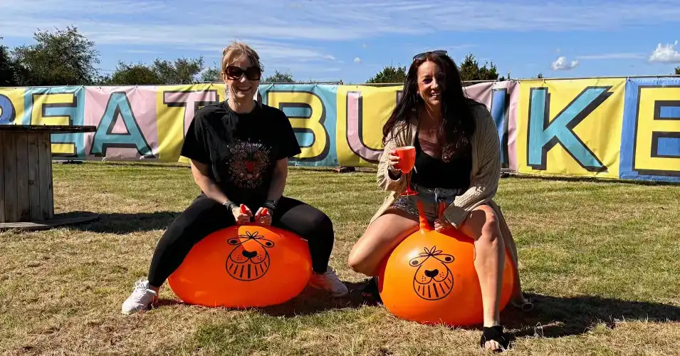 Two women sitting on orange space hoppers with drinks in hand, smiling in front of a colorful Beat Bunker banner on a sunny day.