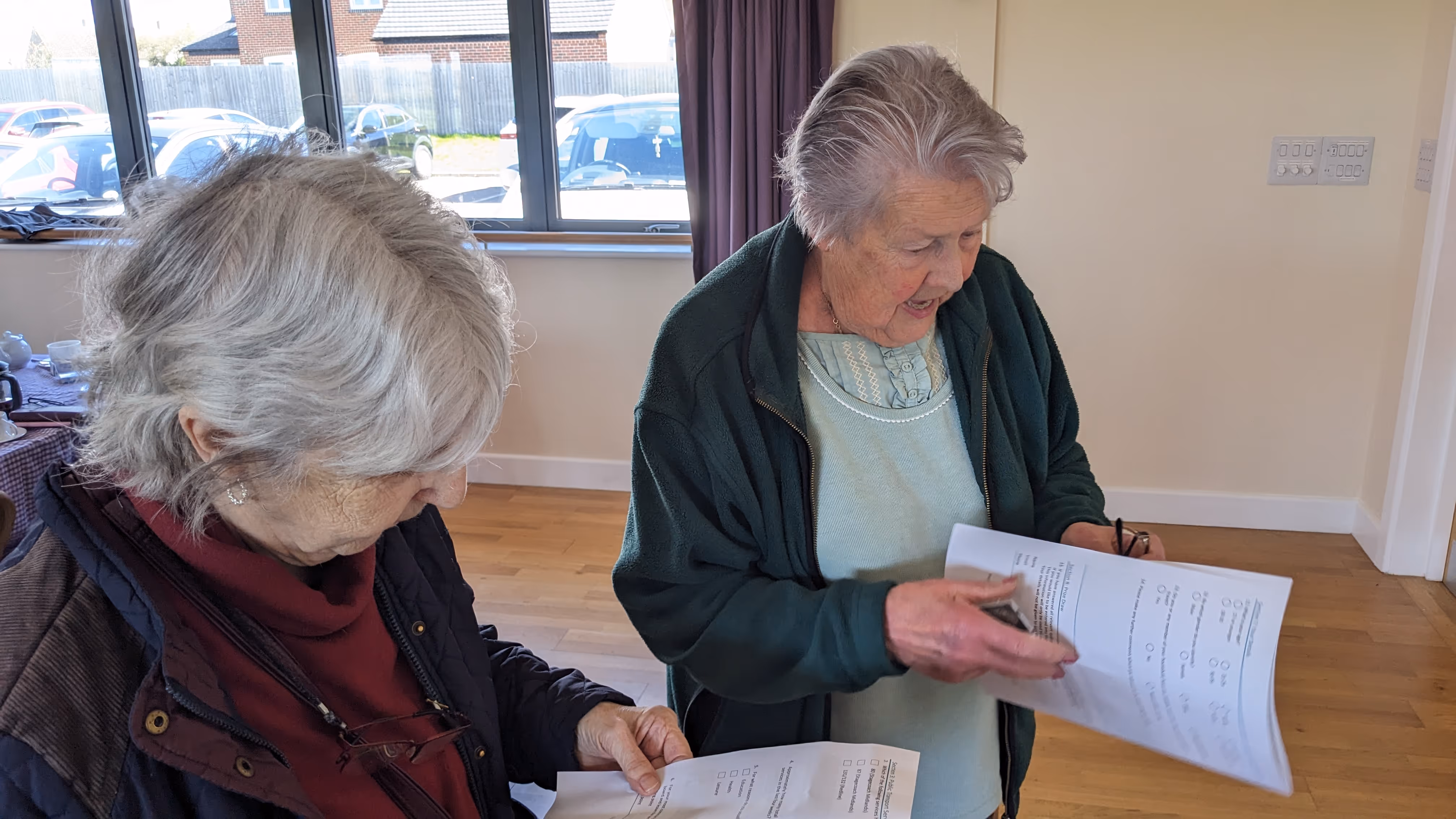 Two elderly women indoors reading and discussing papers, with windows and parked cars behind them.