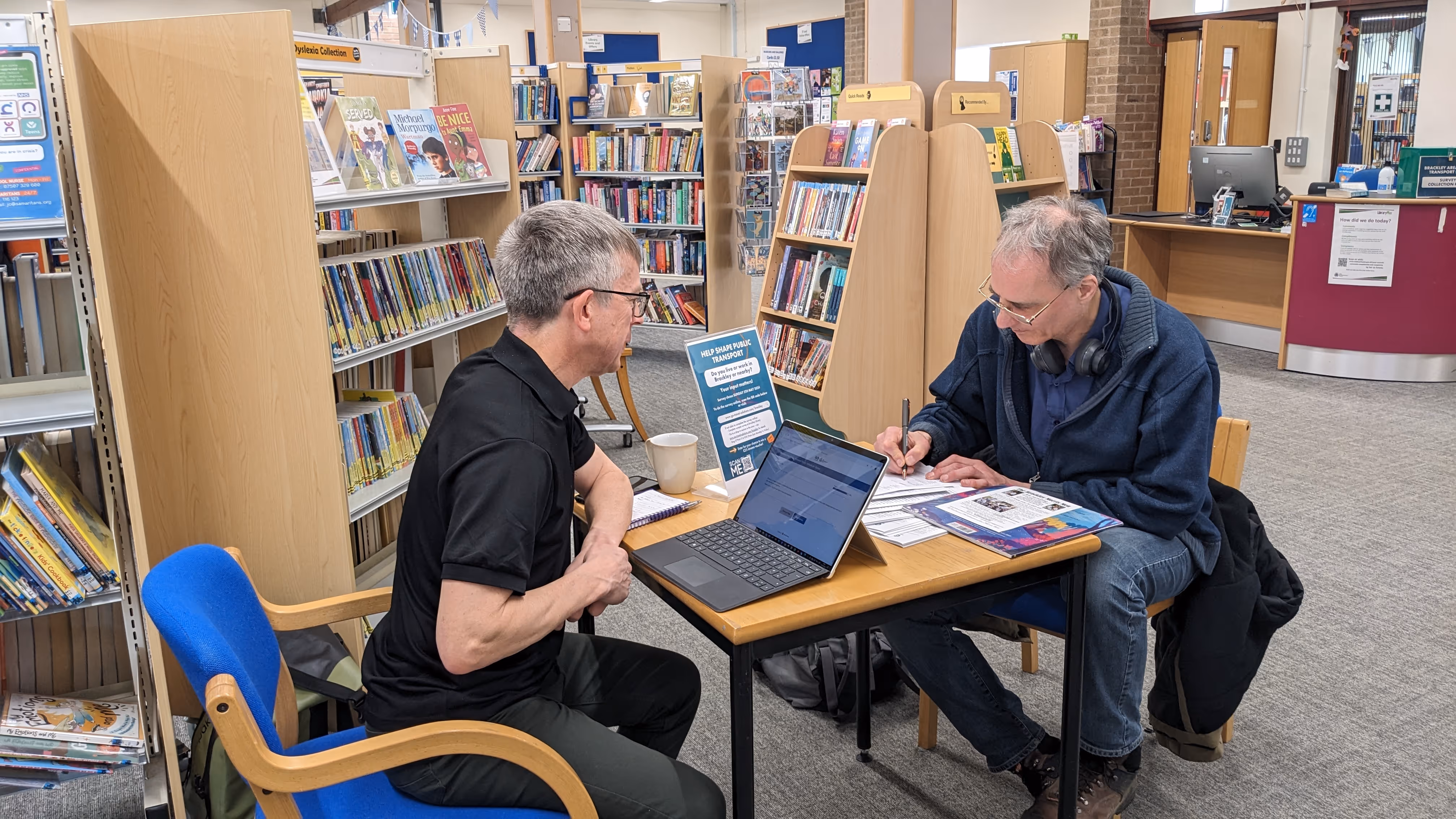 Two men sitting at a library table with laptop and papers, surrounded by bookshelves.