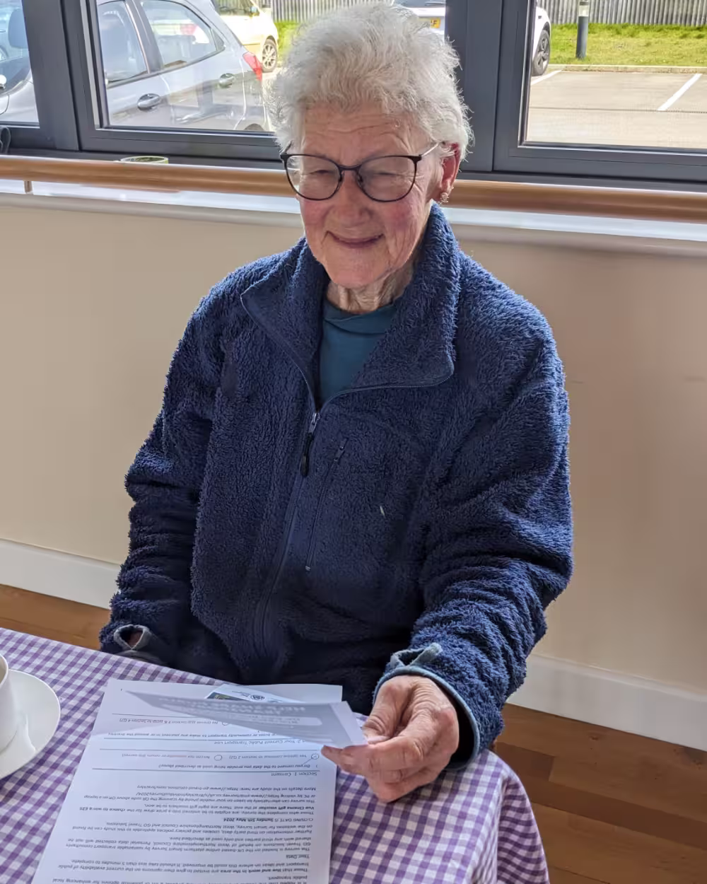 Elderly woman in glasses and fleece jacket sitting at a table with documents and tea cup.
