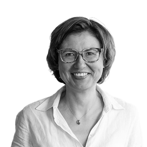 Porträt einer lächelnden Frau mit kurzen Haaren, Brille und weißer Bluse vor weißem Hintergrund.