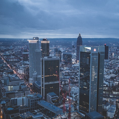 Blick auf die Skyline von Frankfurt am Main mit mehreren beleuchteten Hochhäusern bei wolkenverhangenem Himmel in der Dämmerung.
