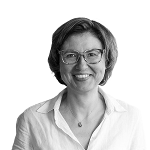 Porträt einer lächelnden Frau mit kurzen Haaren, Brille und weißem Hemd vor weißem Hintergrund.