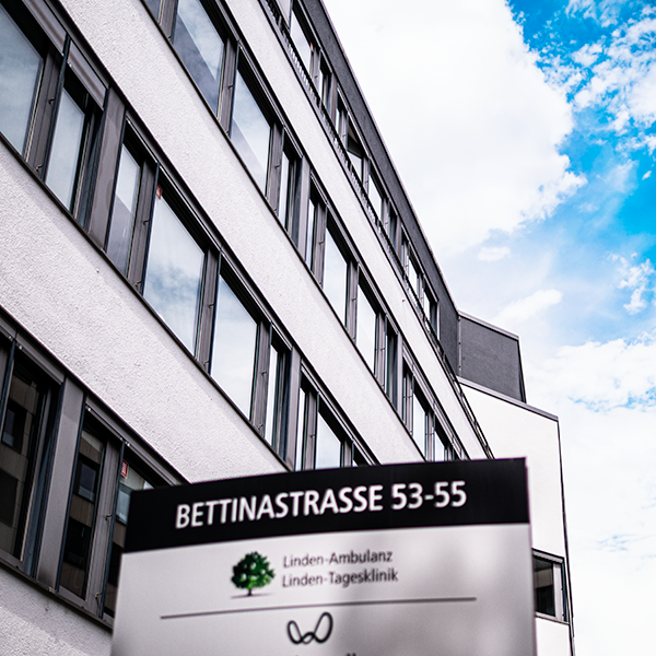 Blick auf ein modernes Büro- oder Klinikgebäude mit vielen Fenstern unter blauem Himmel und einem Schild mit der Adresse Bettinastrasse 53-55 und der Aufschrift Linden-Ambulanz Linden-Tagesklinik.