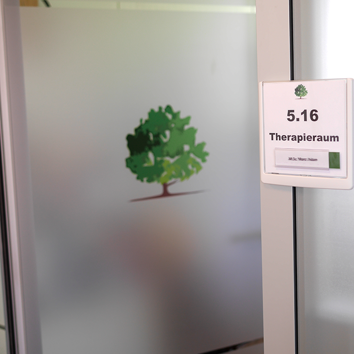 Tür mit mattiertem Glas und grünem Baumlogo sowie Schild mit der Nummer 5.16 und der Aufschrift ‚Therapieraum‘.