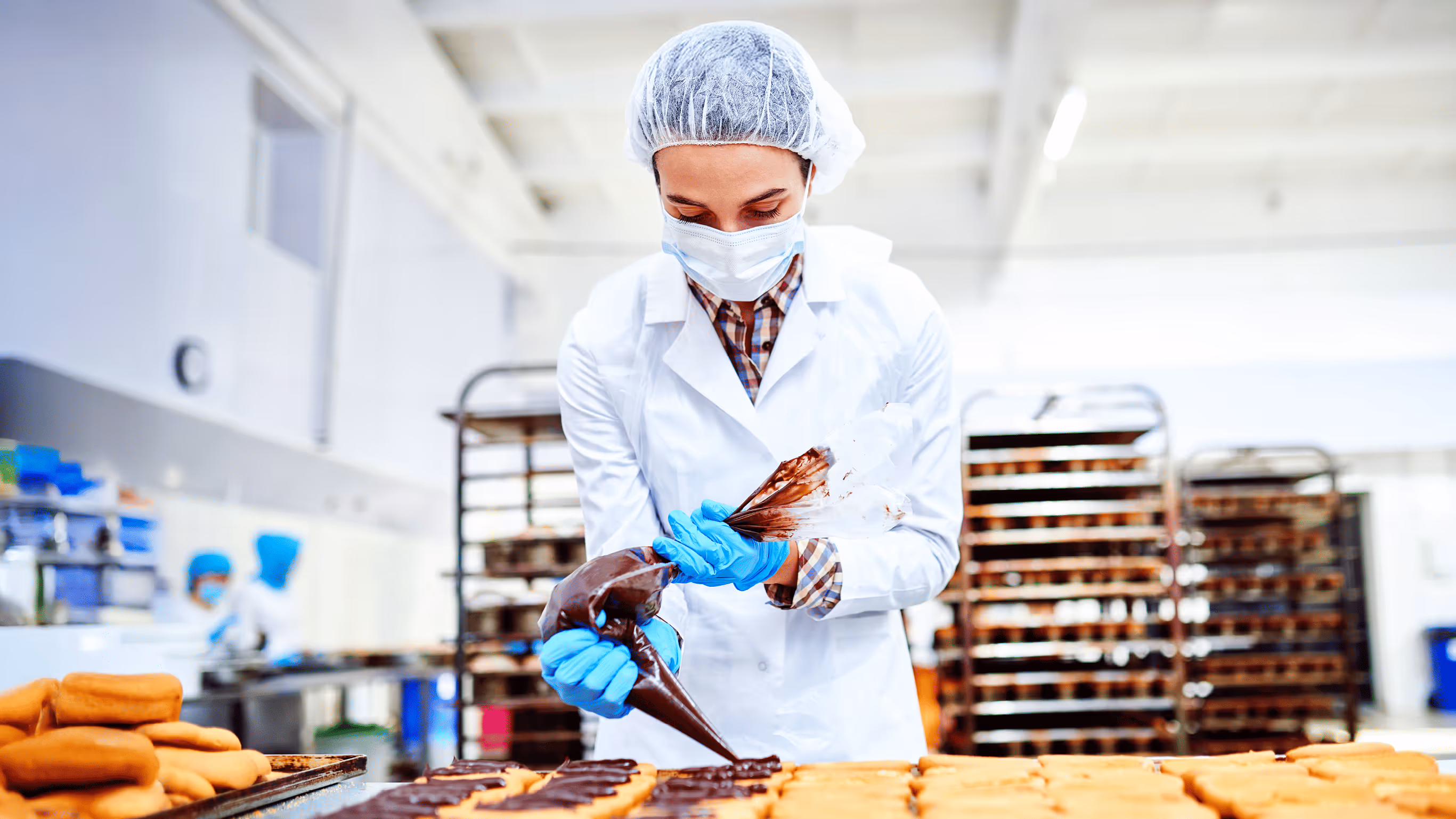 Baker in protective gear decorating pastries with chocolate in food factory