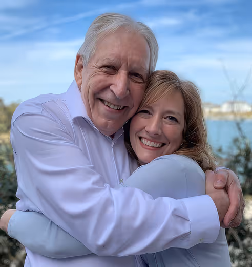 Smiling elderly man and woman hugging outdoors with a lake and houses in the background.