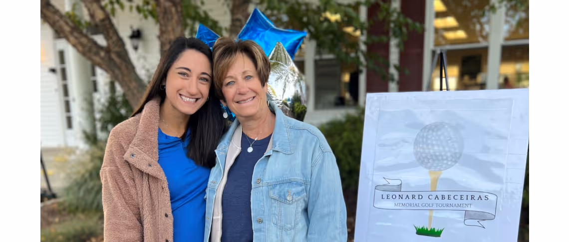 Two smiling women standing outdoors near a sign for the Leonard Cabeceiras Memorial Golf Tournament, with blue and silver balloons in the background.