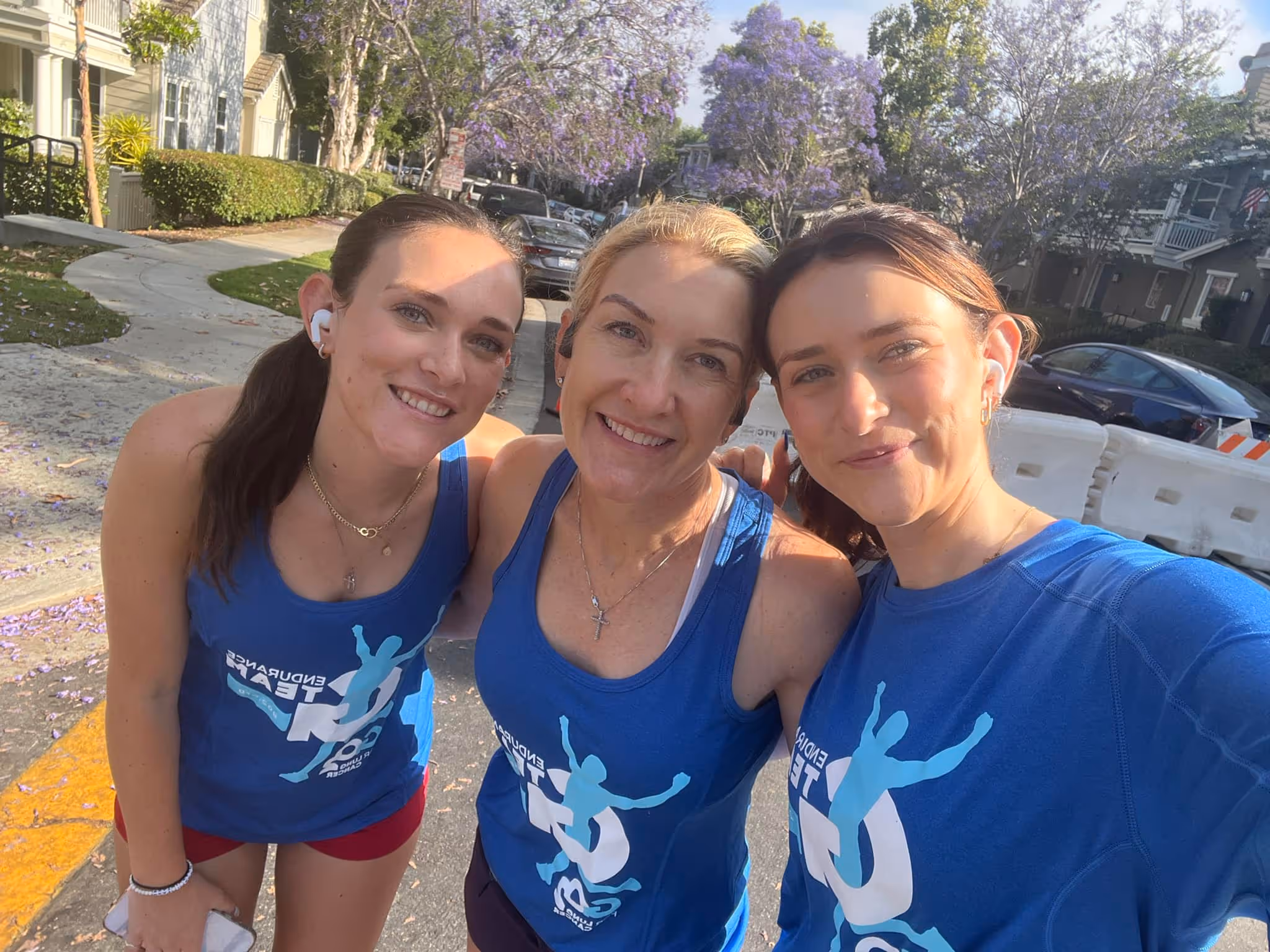 Three women in blue running shirts smiling and posing for a selfie outdoors on a sunny day.