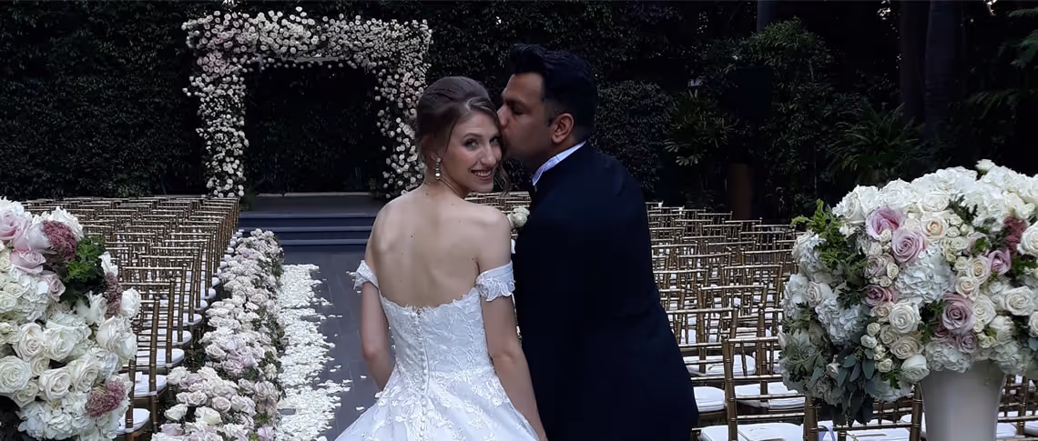 Bride in white dress smiling at the camera and groom in black suit kissing her cheek at an outdoor wedding ceremony with floral arch and decorated aisle.