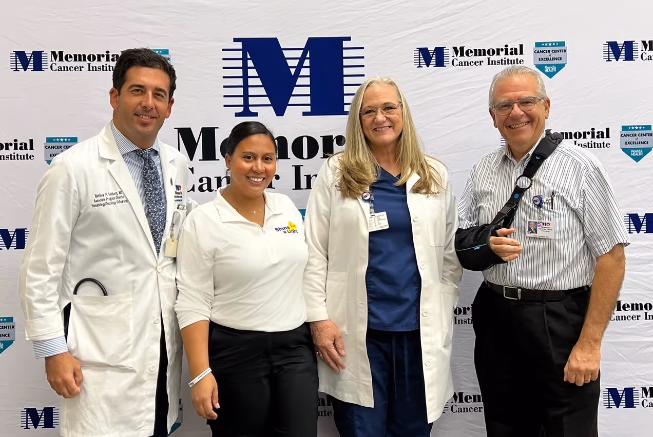 Four medical professionals smiling and posing in front of a Memorial Cancer Institute backdrop, two wearing white coats and one in a Shine a Light polo shirt.
