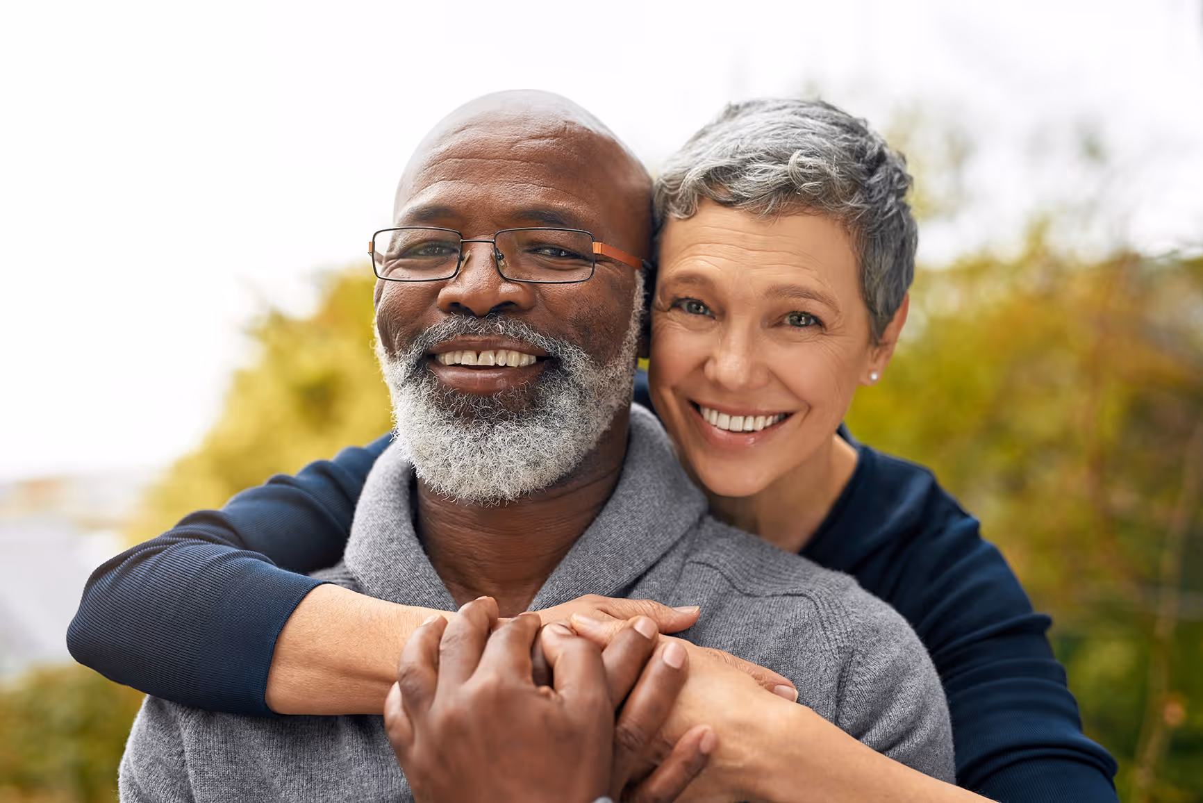Smiling middle-aged couple embracing outdoors with greenery in the background.