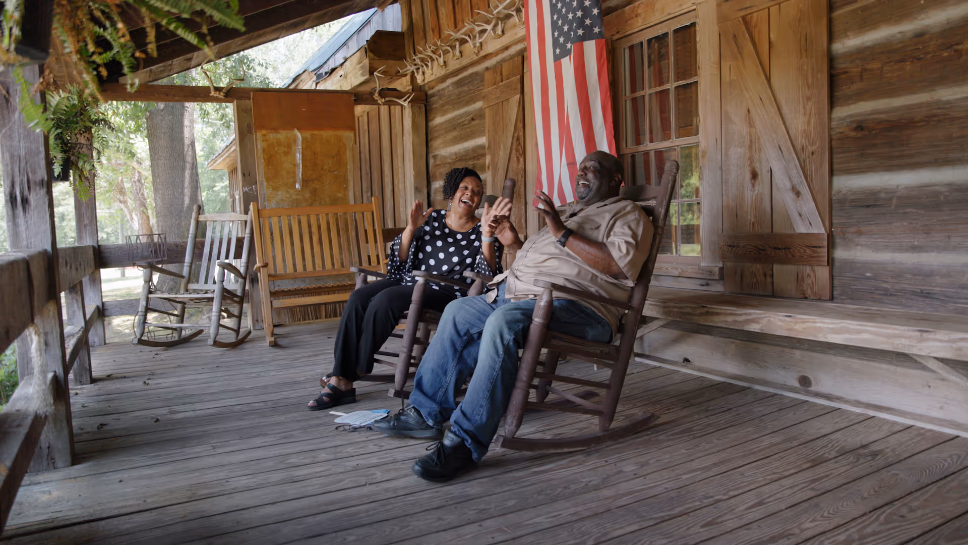 An elderly couple laughing and talking while sitting on rocking chairs on a wooden porch with an American flag displayed on the wall behind them.