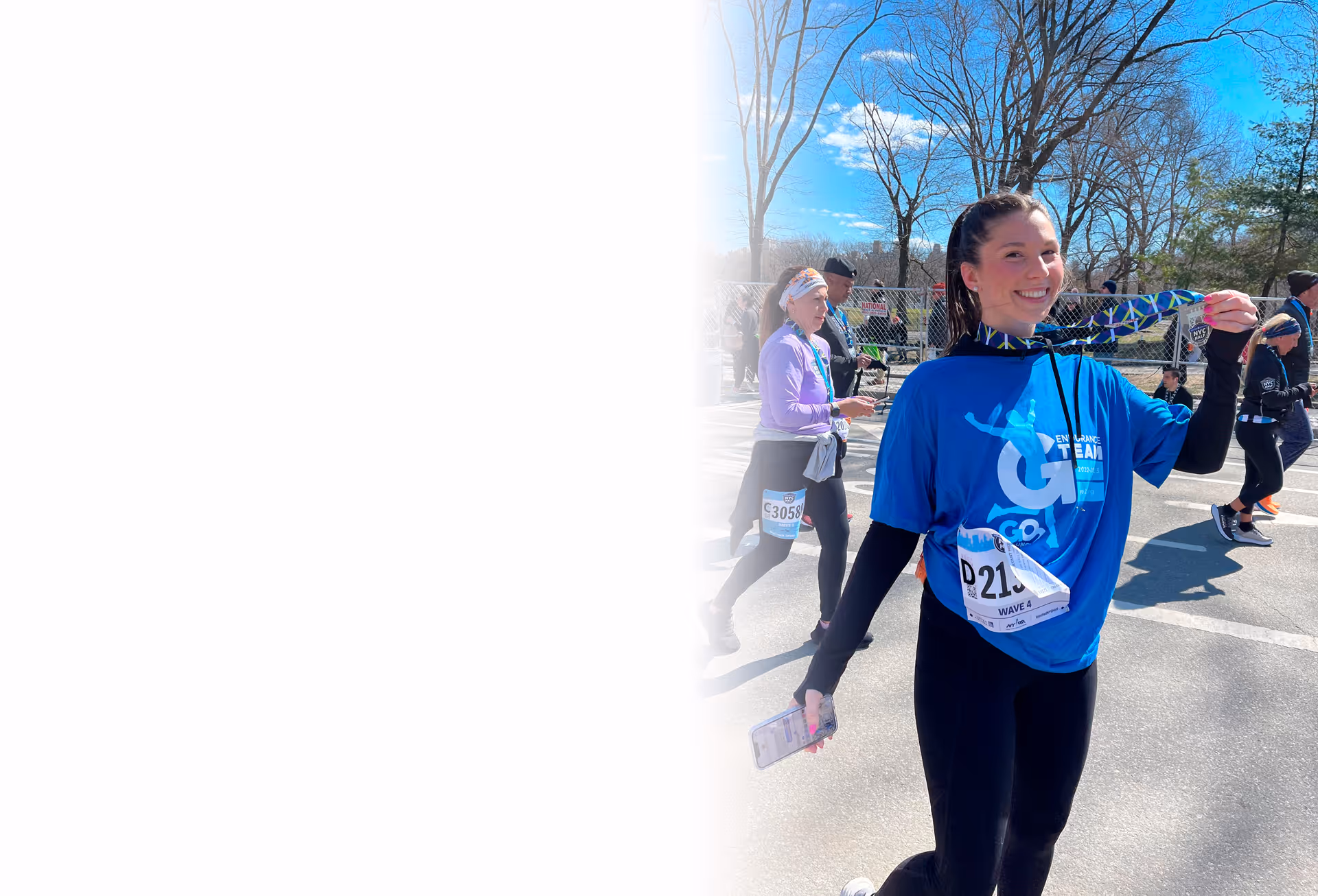 Smiling female runner holding a medal and phone, wearing blue marathon shirt and race bib, with other runners in background on a sunny day.