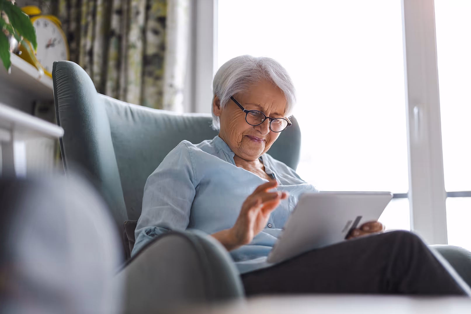 Elderly woman with glasses sitting in a comfortable chair using a tablet device.