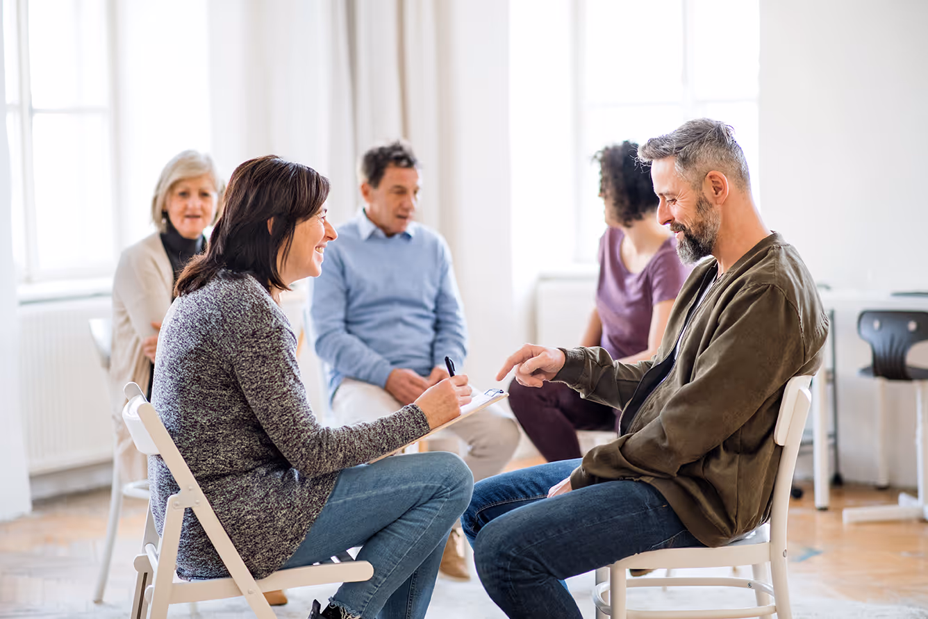 A group therapy session with two people in the foreground engaged in conversation and others seated in the background.
