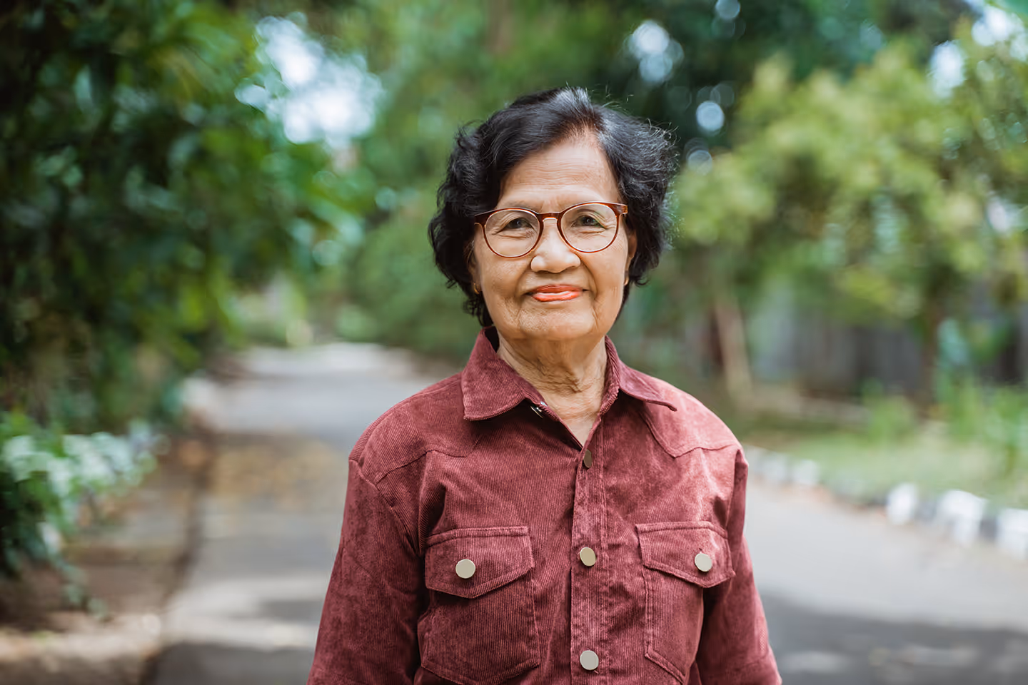 Elderly woman with short curly hair and glasses wearing a maroon jacket standing outdoors on a blurred tree-lined path.