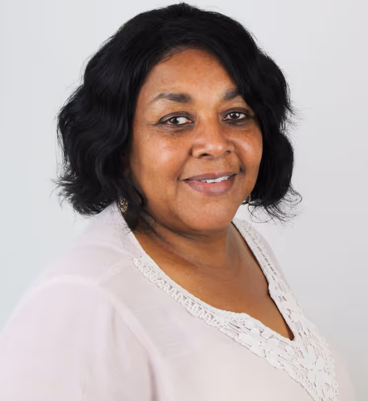 Smiling middle-aged woman with curly black hair wearing a light pink top with lace detail against a plain white background.