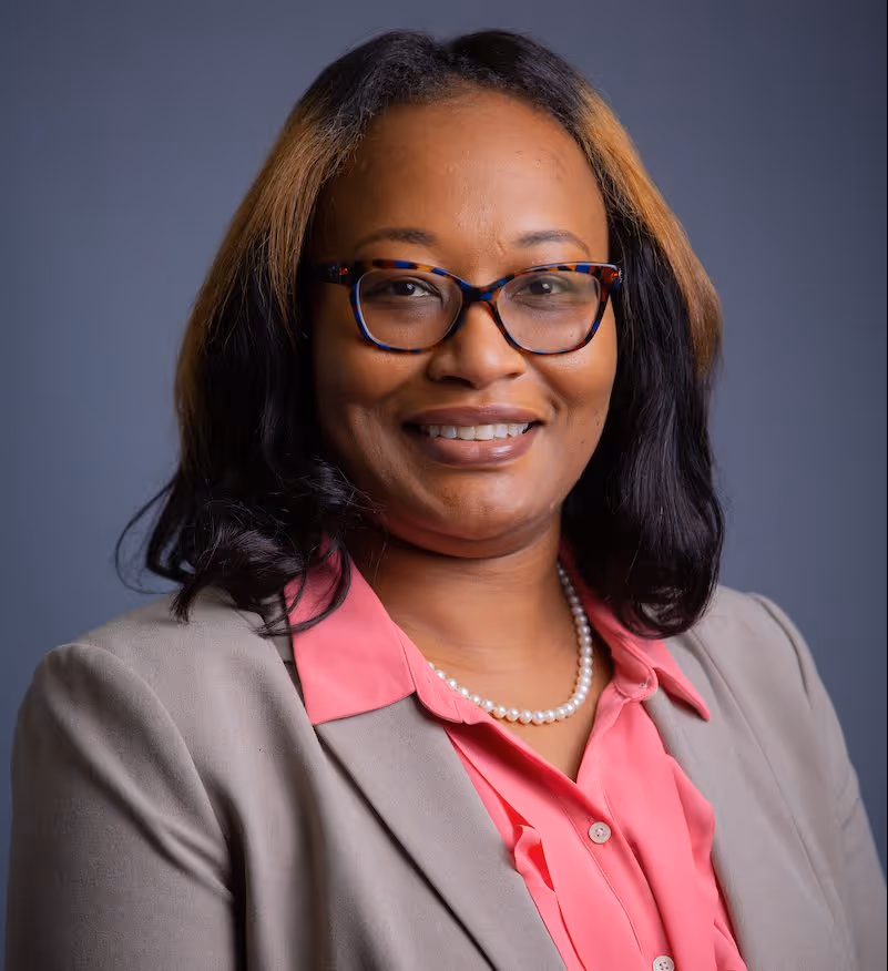 Smiling woman with black and brown hair wearing tortoiseshell glasses, pink blouse, gray blazer, and a pearl necklace against a gray background.