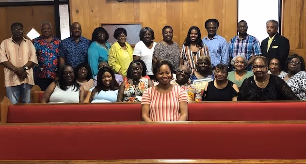 Group of 21 adults posing and smiling inside a room with wood-paneled walls, with some seated on church pews and others standing behind them.