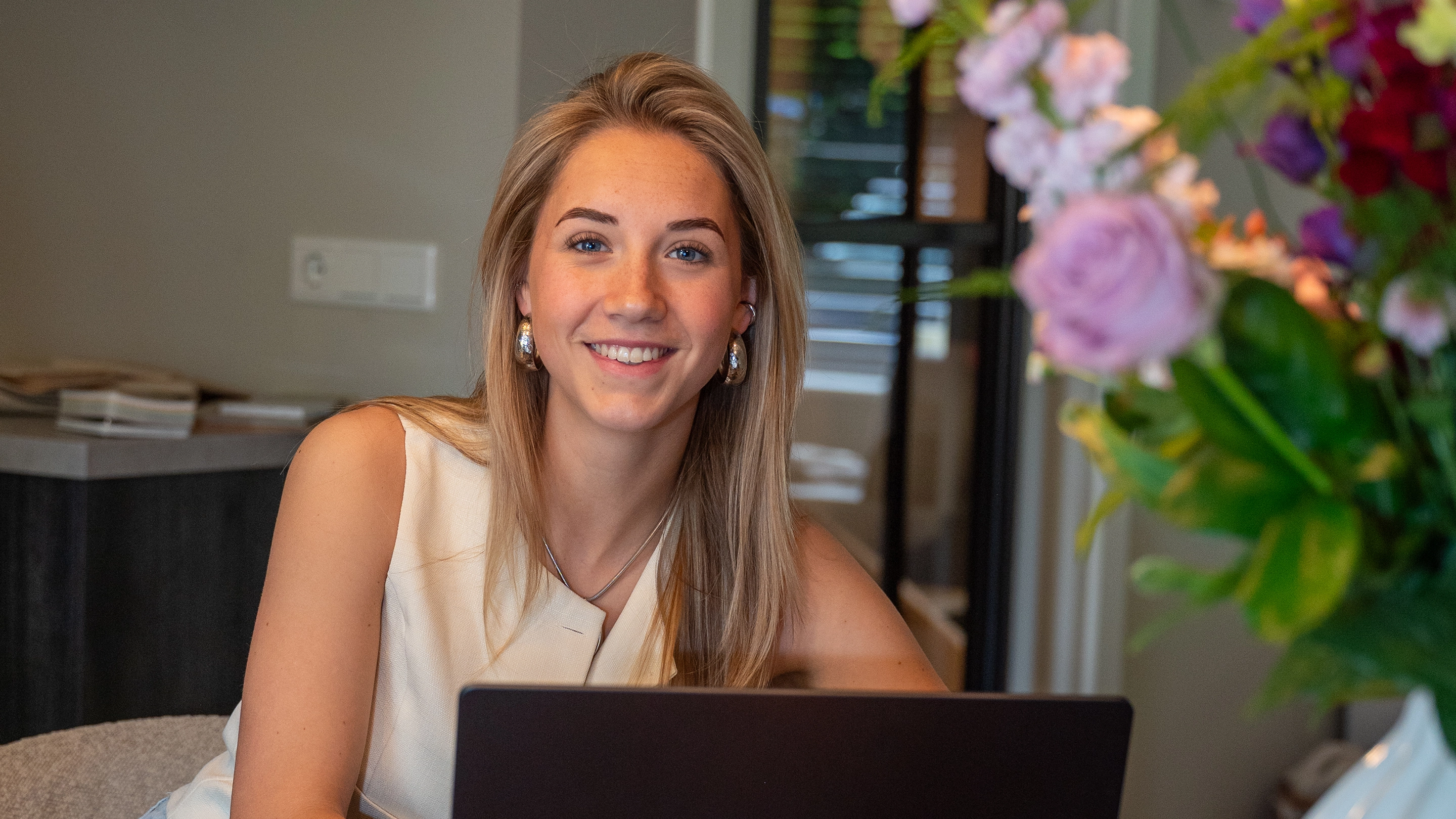 Interieurontwerpster Anouk glimlacht achter haar laptop tijdens een persoonlijk adviesgesprek, met bloemen op de voorgrond.