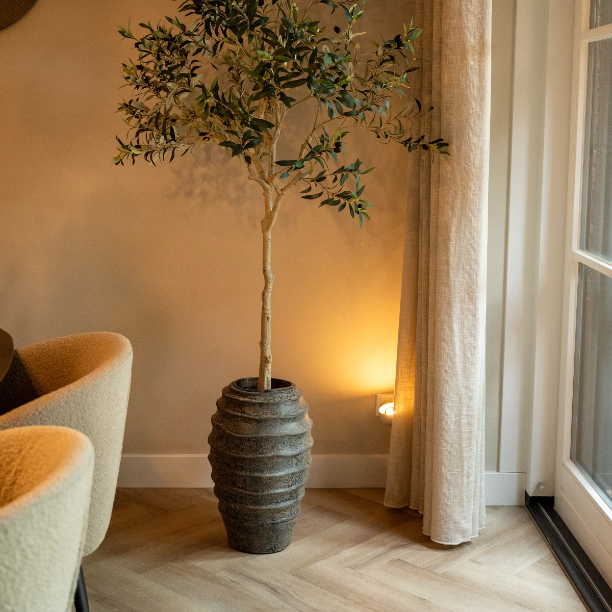 Stijlvol interieurdetail met olijfboom in grote keramische pot naast linnen gordijnen en zachte fauteuil, styling door Nouk Interiors.