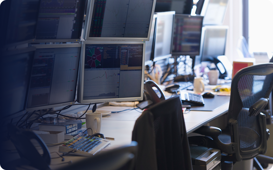 Office workspace with multiple computer monitors displaying charts and data, ergonomic chairs, keyboards, and coffee mugs on desks.