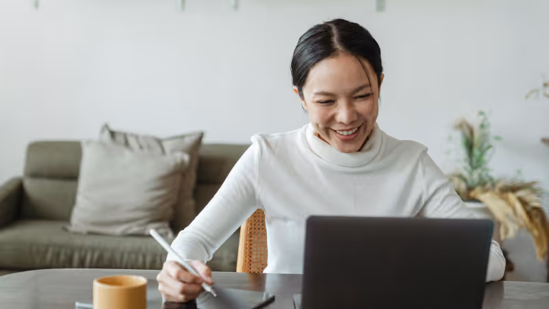 Smiling woman using a stylus tablet and laptop at a table in a cozy living room.