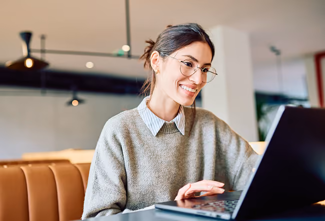 Enterprise Video Platform Overview Smiling woman wearing glasses and a gray sweater sitting in a modern office space, looking at a computer screen.