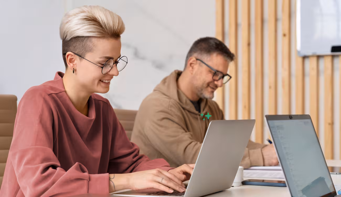 Zwei Kolleg:innen arbeiten nebeneinander an ihren Laptops, einer macht sich handschriftliche Notizen.