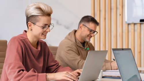 Zwei Kolleg:innen arbeiten nebeneinander an ihren Laptops, einer macht sich handschriftliche Notizen.