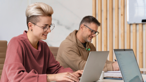 Zwei Kolleg:innen arbeiten nebeneinander an ihren Laptops, einer macht sich handschriftliche Notizen.