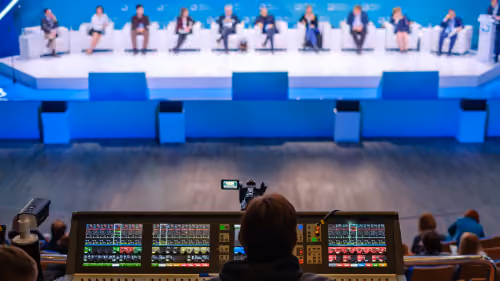 One-Time Event: Livestreaming für einmalige Veranstaltungen | movingimage Kamera und Tonpult filmen eine Podiumsdiskussion mit elf Teilnehmenden auf einer Konferenzbühne