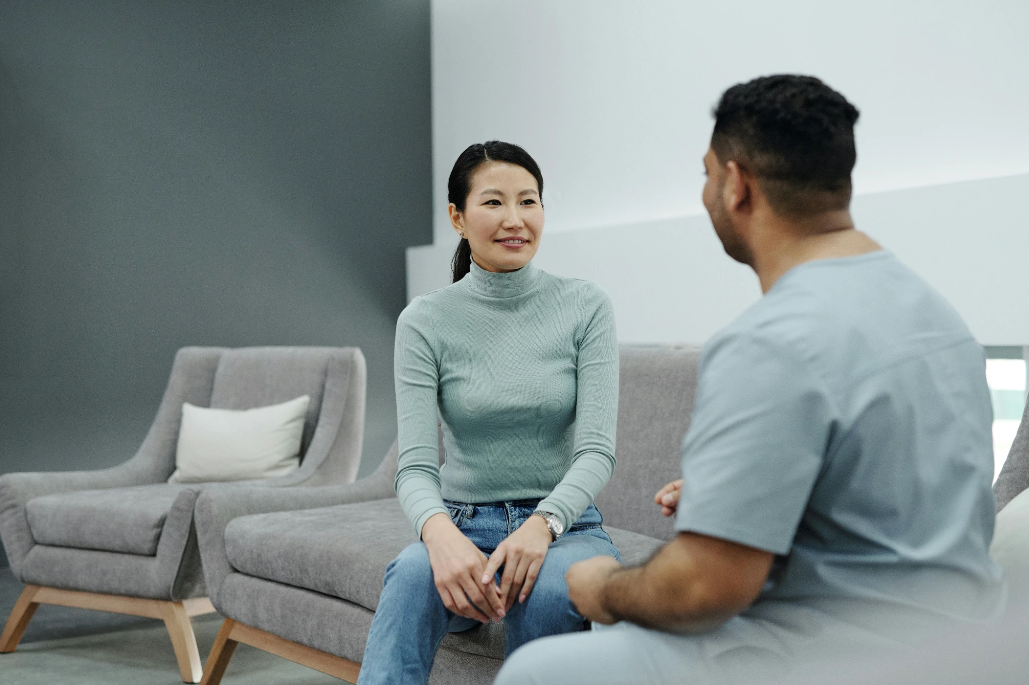 Woman in a green turtleneck and jeans sitting on a gray couch talking to a man in medical scrubs.