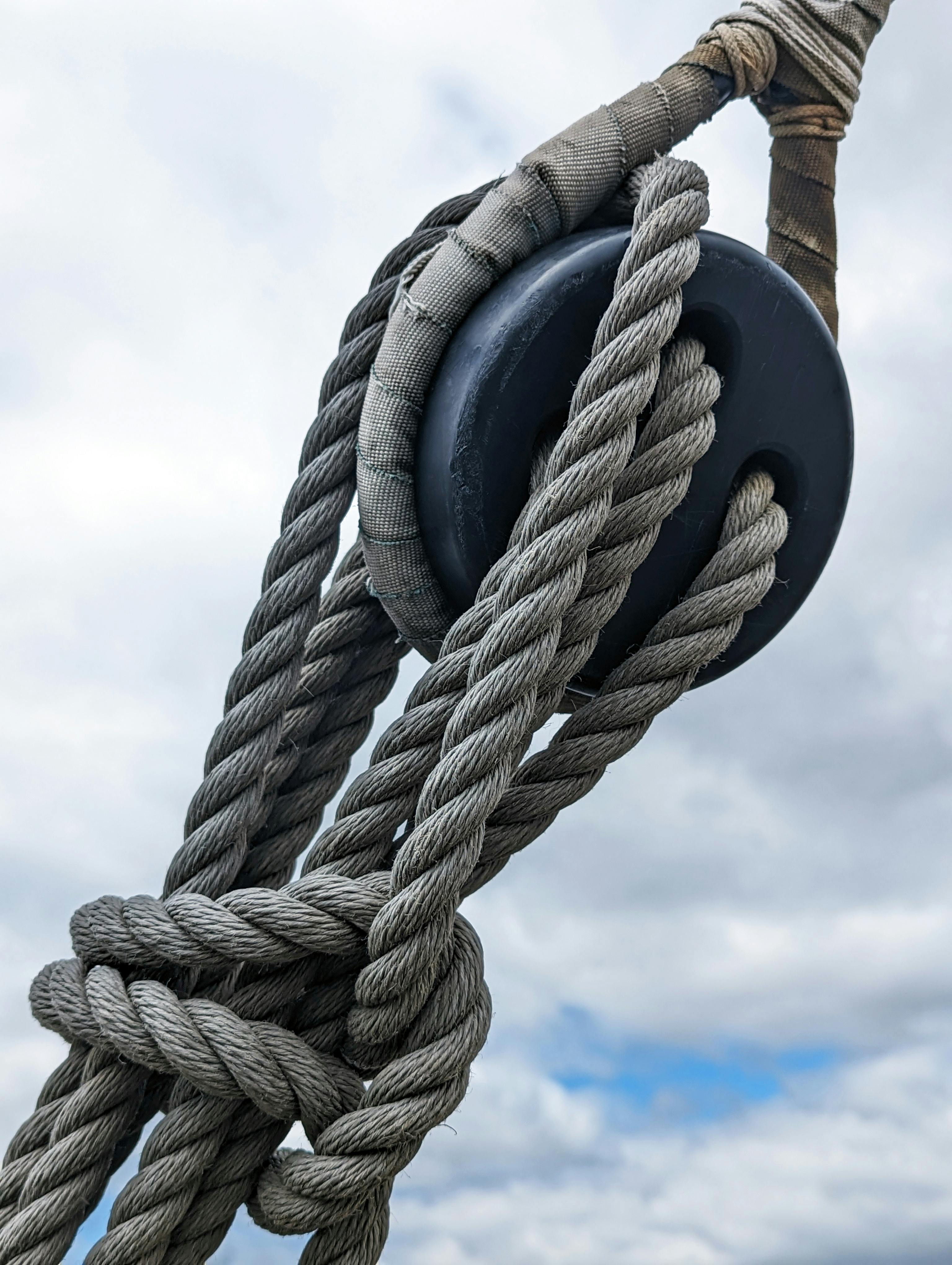 Close-up of gray ropes threaded through a black pulley against a cloudy sky.