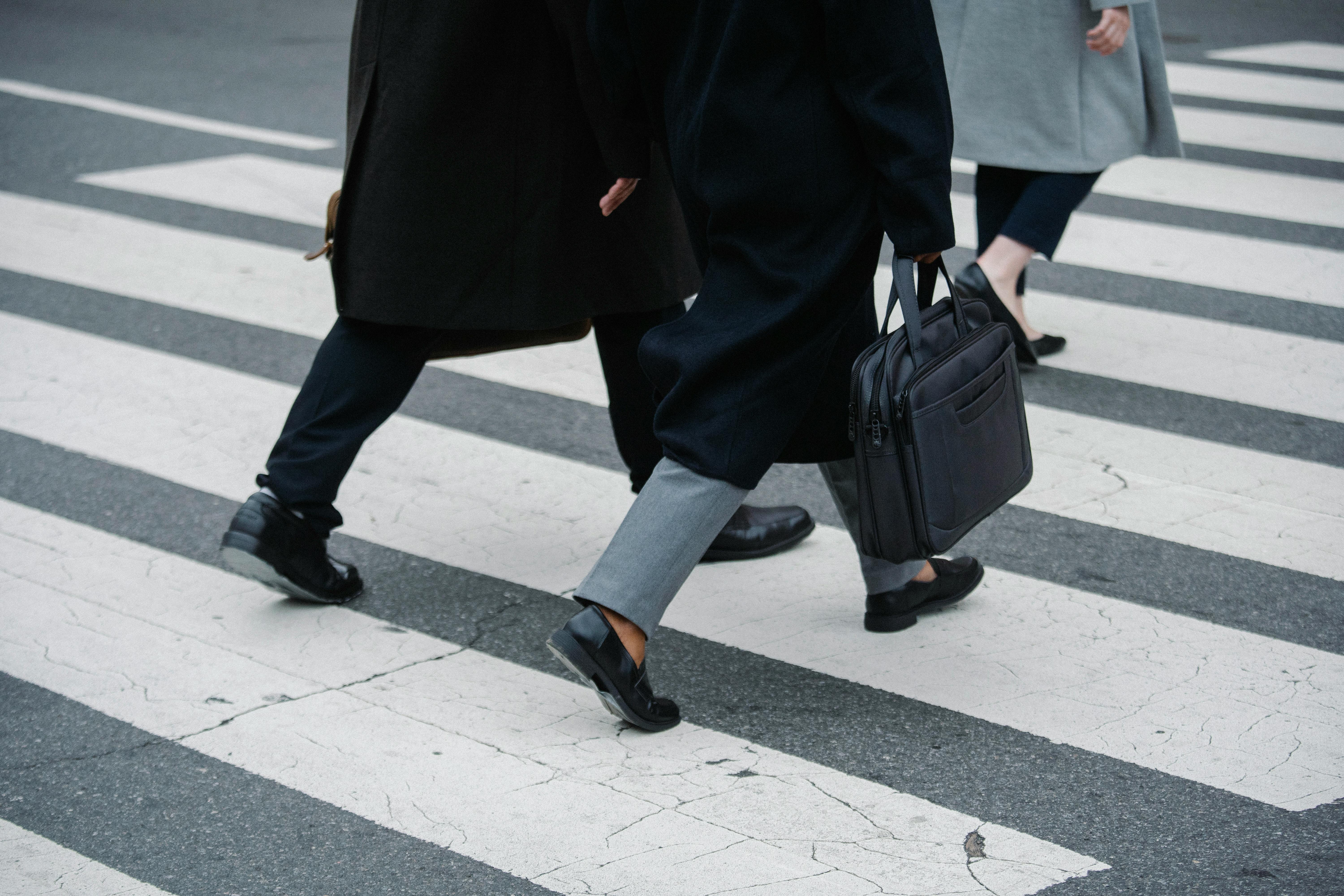 Three people walking across a city crosswalk wearing coats and black dress shoes, one carrying a black briefcase.