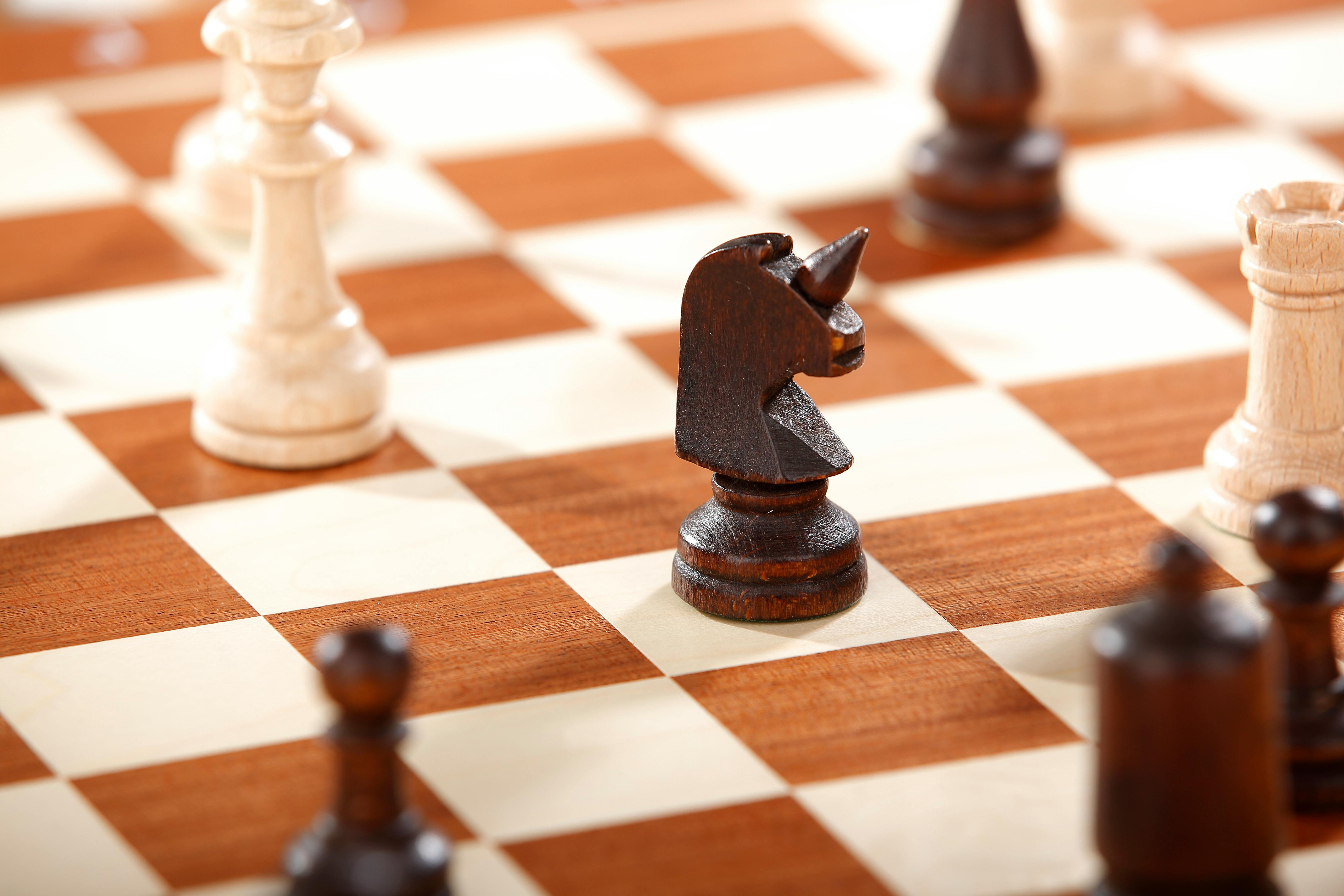 Close-up of a dark wooden knight chess piece on a chessboard with other blurred pieces in the background.